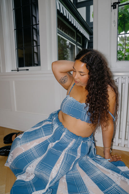 A woman with long curly dark hair sits on a wooden floor indoors, wearing a blue and white plaid two-piece outfit consisting of a cropped tank top and a matching wide skirt.